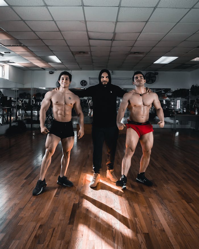 Two bodybuilders with their trainer posing confidently in a gym setting.