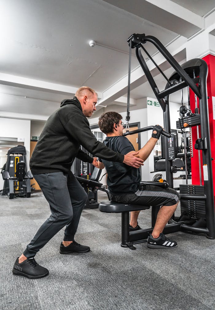 A personal trainer guides a client performing a workout using a gym machine, emphasizing correct form and technique.