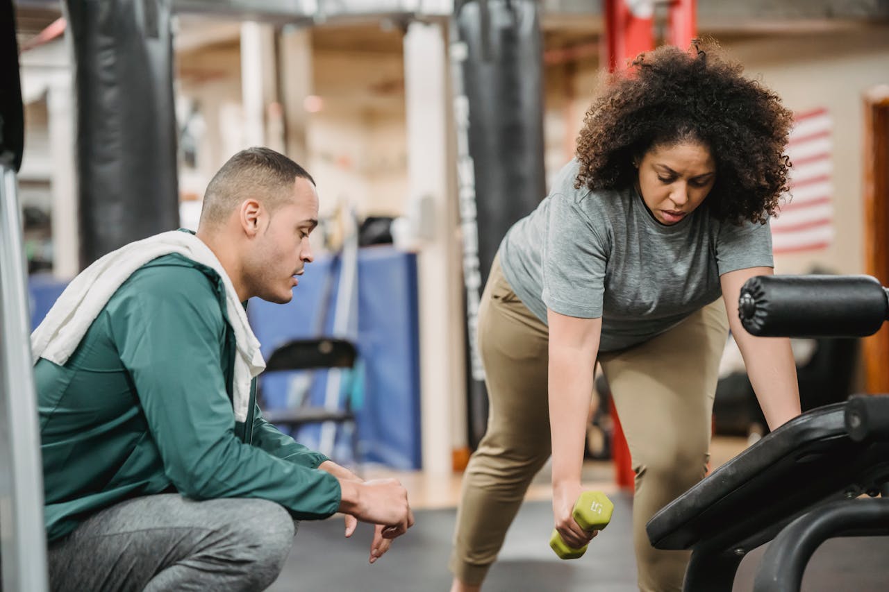 A personal trainer guides a woman lifting dumbbells in a gym setting.